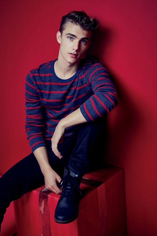 Young adult male model poses indoors against a red background wearing a striped sweater and sitting on a red wrapped gift box.