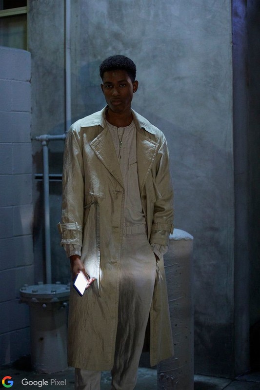 Black man wearing a long beige trench coat holding a glowing smartphone in an urban alleyway at night.