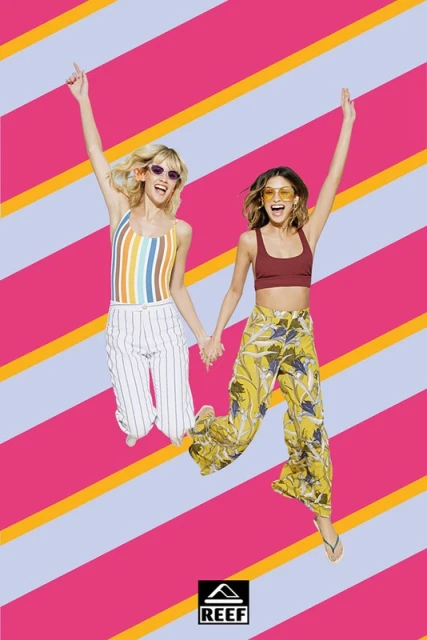 Two adult females jumping in the air with arms raised, wearing sunglasses, tops, and wide-leg pants in front of a striped background.
