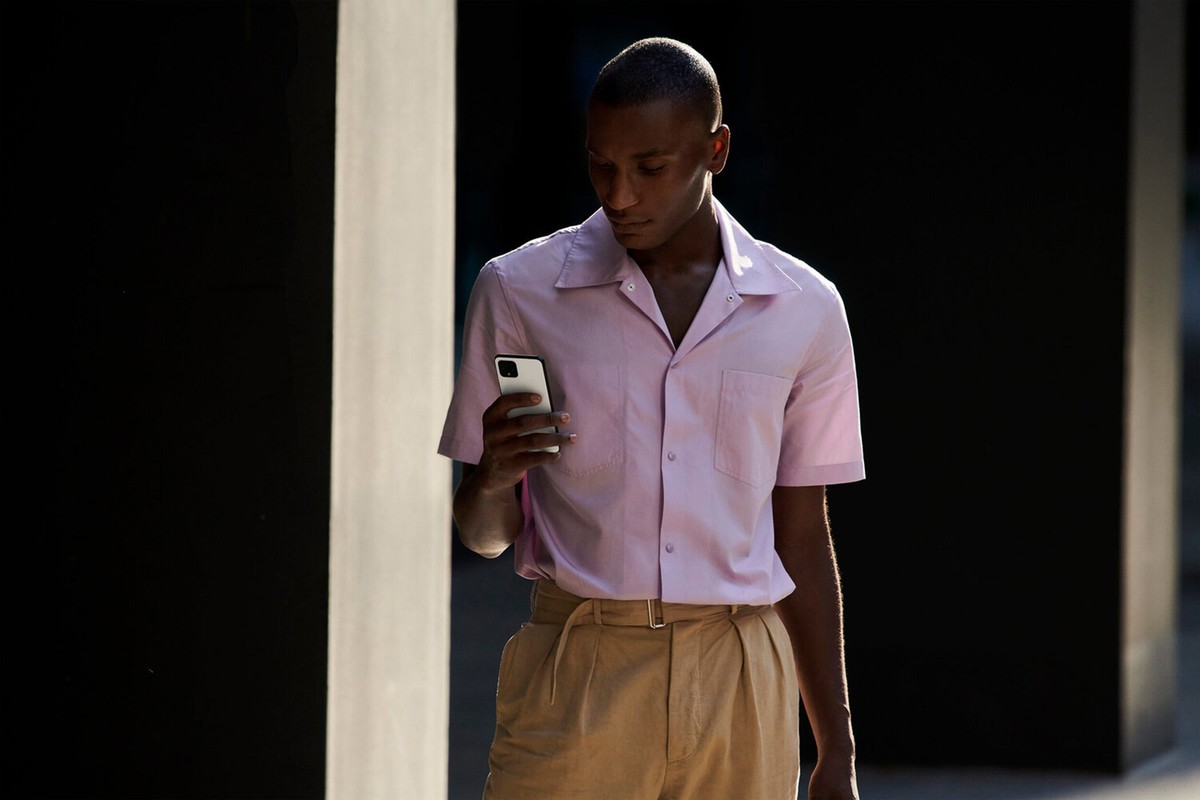 An African American man in a pink short-sleeved shirt and tan trousers looks at a white smartphone in an outdoor urban setting.
