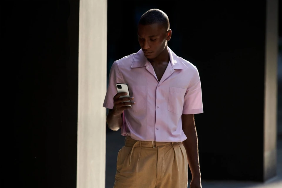 An African American man in a pink short-sleeved shirt and tan trousers looks at a white smartphone in an outdoor urban setting.