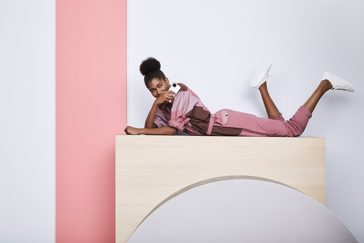 Adult female lies on a light-brown wood structure, holds a white smartphone, wears brown and rose-color jumpsuit and white sneakers, indoor setting.