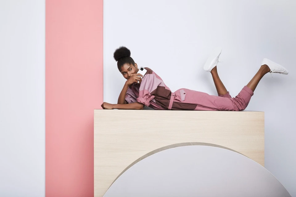 Adult female lies on a light-brown wood structure, holds a white smartphone, wears brown and rose-color jumpsuit and white sneakers, indoor setting.