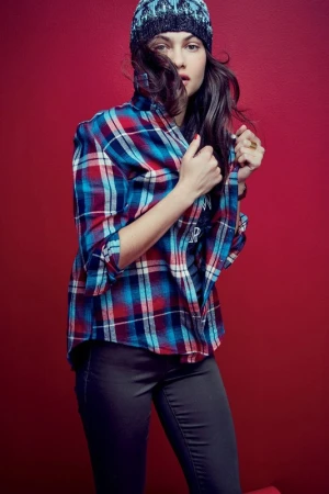 Woman wearing a blue and red plaid shirt and patterned beanie posing against a bold red background