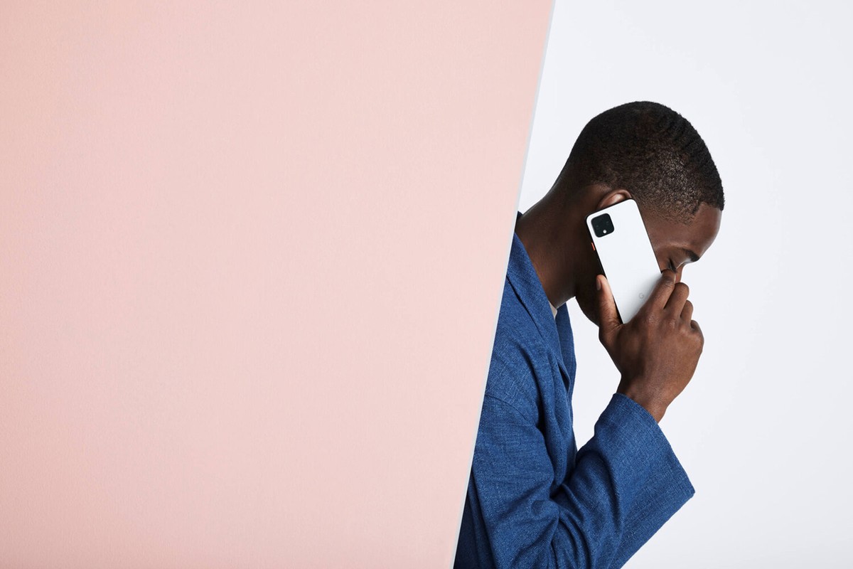 A dark-skinned adult male wearing a blue jacket holds a white smartphone to his ear in a studio setting with a pink and white backdrop.