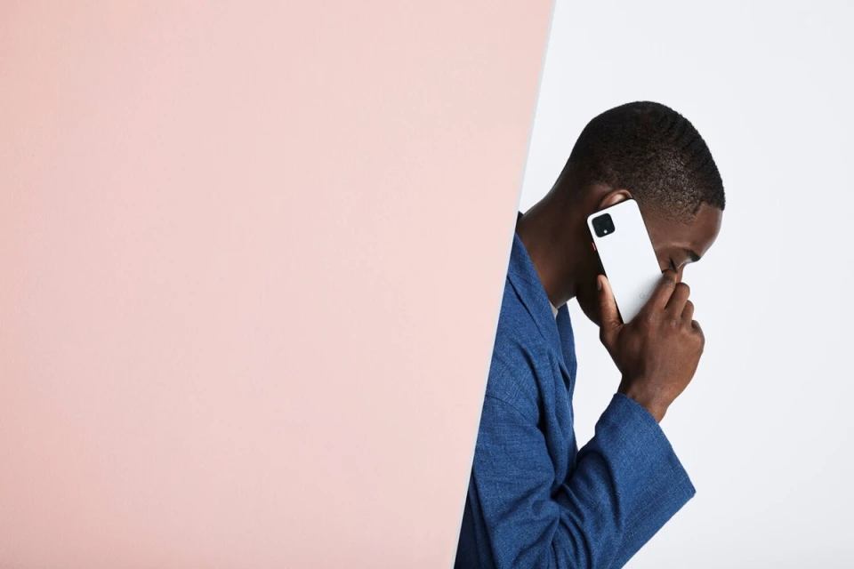 A dark-skinned adult male wearing a blue jacket holds a white smartphone to his ear in a studio setting with a pink and white backdrop.