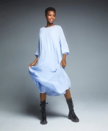 Black woman in a light blue midi dress and black combat boots smiling against a gradient grey studio backdrop.