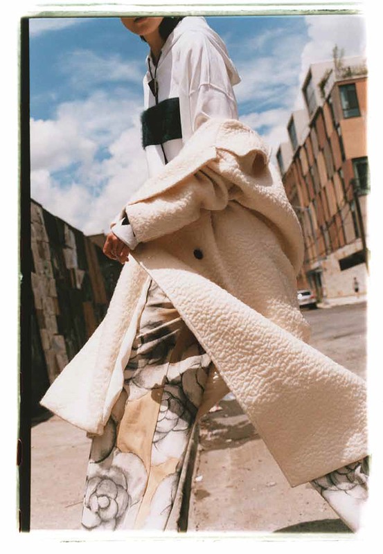 Fashion shot of a person in a coat and patterned pants walking in an urban setting on a sunny day with a blue, cloudy sky.