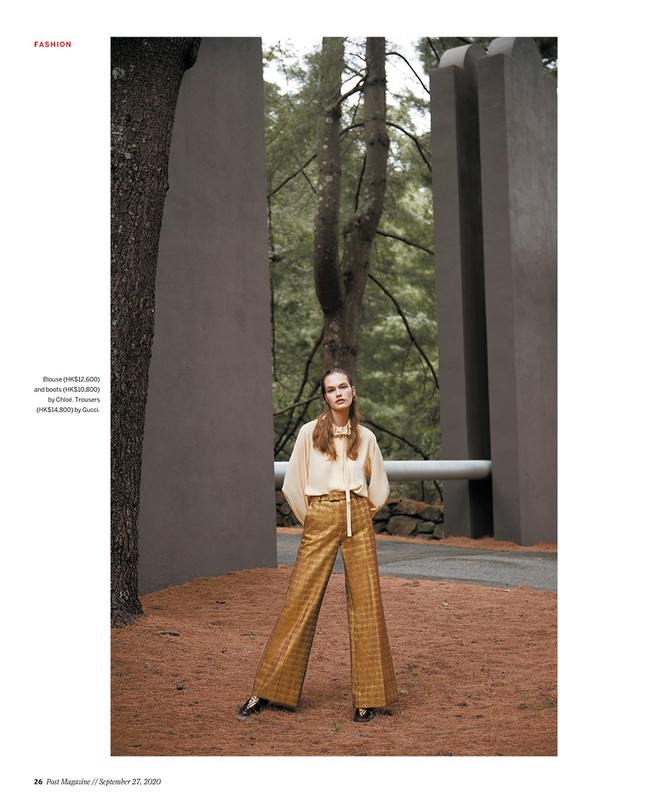 Fashion editorial shot of a woman in gold patterned pants and a cream blouse, posing in a natural outdoor environment with trees.