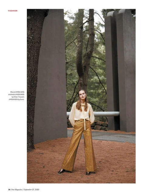 Fashion editorial shot of a woman in gold patterned pants and a cream blouse, posing in a natural outdoor environment with trees.