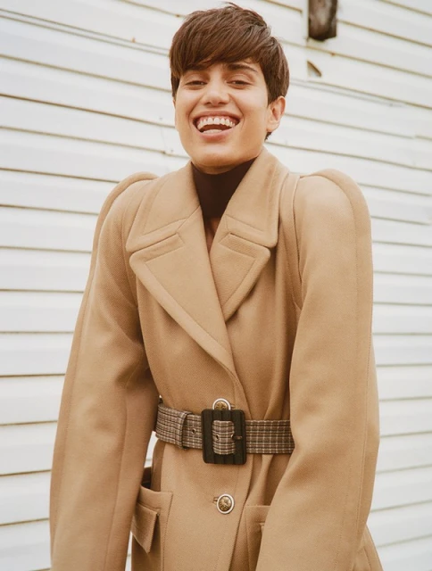 Smiling woman with short hair, wearing a brown turtleneck and tan overcoat with a patterned belt, posing against a white wall.
