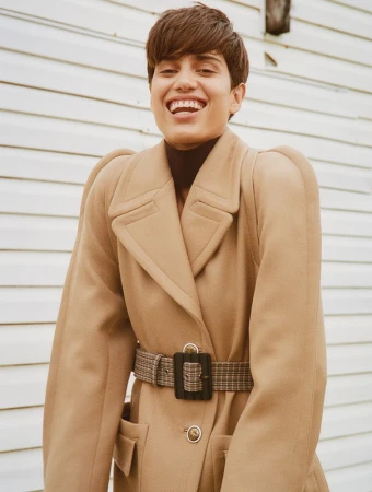 Smiling woman with short hair, wearing a brown turtleneck and tan overcoat with a patterned belt, posing against a white wall.