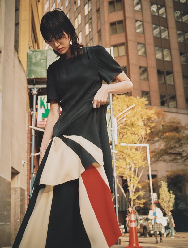 Woman in a black dress with white and red color blocks poses on a city sidewalk with tall buildings and traffic lights in the background.
