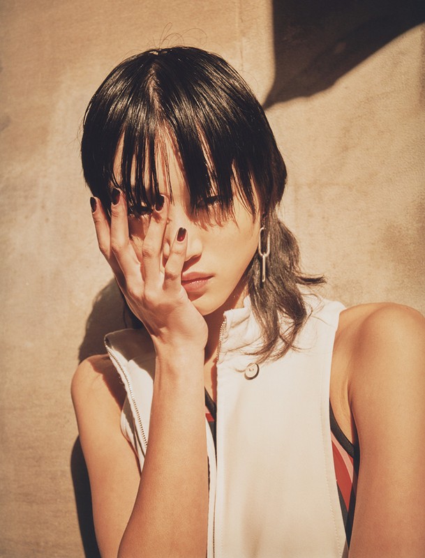 Portrait of a young woman with dark hair covering one eye, wearing a cream-colored vest, with natural light and a beige background.