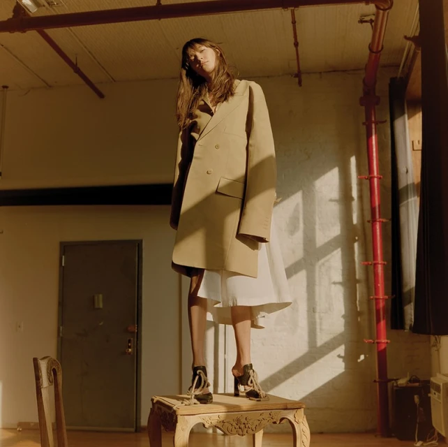 A fashionable young woman in a tan coat and platform heels stands on a decorative table in a room with warm, natural lighting.