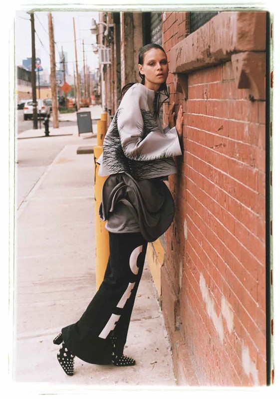 A fashion shot of a woman in black and silver clothing leaning against a brick wall in an urban outdoor setting, with a sidewalk.