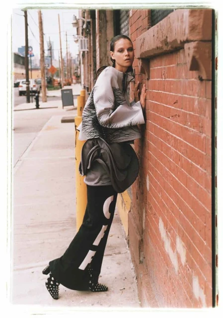 A fashion shot of a woman in black and silver clothing leaning against a brick wall in an urban outdoor setting, with a sidewalk.