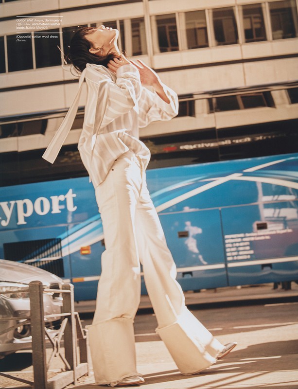 Fashion model poses in white outfit with blue city bus in background. Upward angle captures woman with building backdrop.
