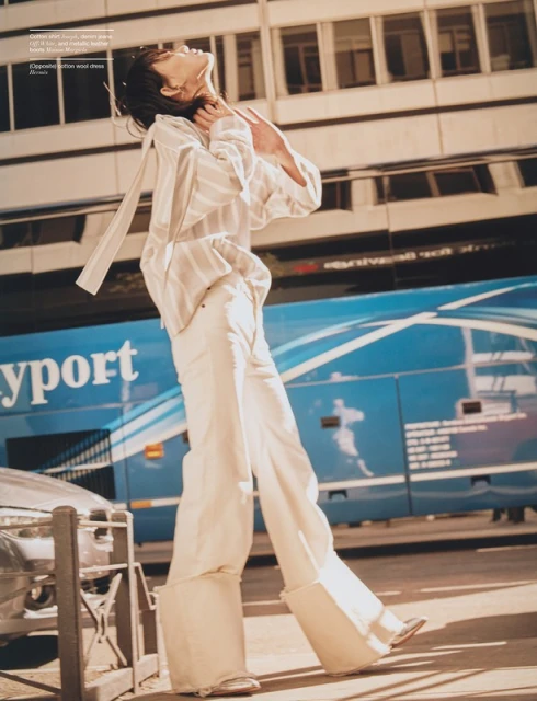 Fashion model poses in white outfit with blue city bus in background. Upward angle captures woman with building backdrop.