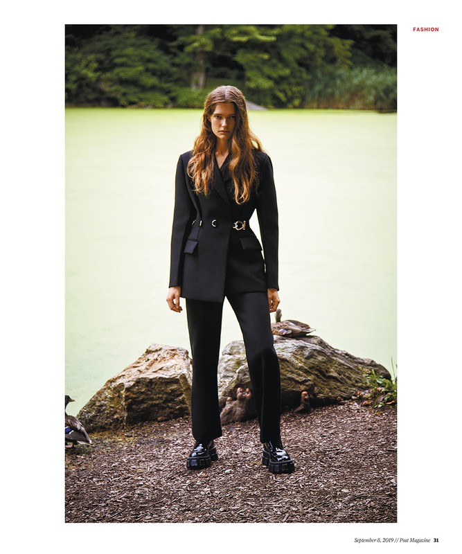 A female fashion model poses in a black blazer and trousers, standing on a path by a green pond and foliage for a fashion editorial.