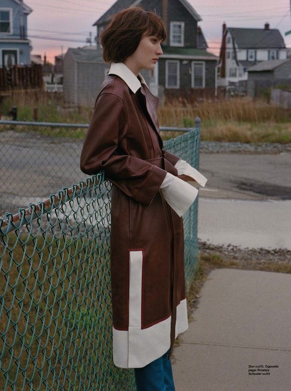 Brunette adult female model wearing brown and cream colored trench coat stands near a chain link fence in an outdoor urban setting.