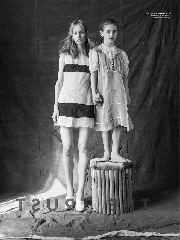 A fashion editorial shot shows two young female models wearing dresses, one barefoot on a stool, against a draped backdrop in monochrome.
