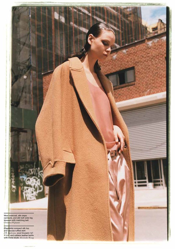Fashion editorial shot of an adult female wearing a tan overcoat and light pink silk suit in a city street setting.