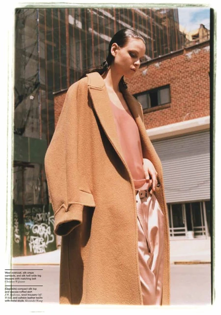 Fashion editorial shot of an adult female wearing a tan overcoat and light pink silk suit in a city street setting.
