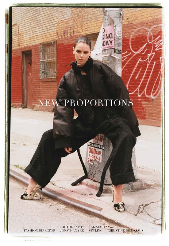 Fashion editorial image of a woman in black outerwear and heels sitting on a metal bar, urban street background with brick buildings and graffiti.