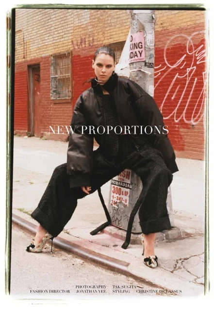 Fashion editorial image of a woman in black outerwear and heels sitting on a metal bar, urban street background with brick buildings and graffiti.