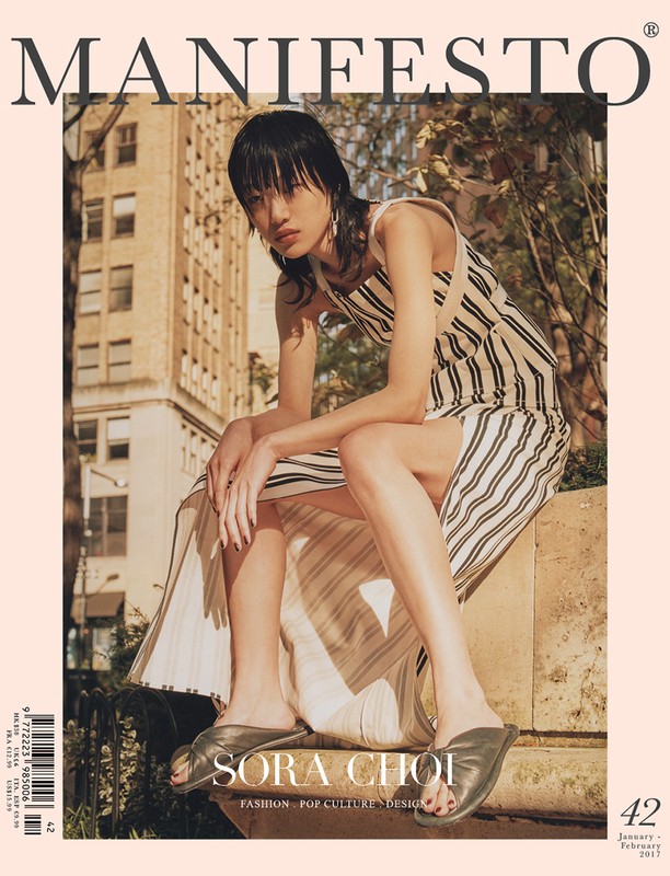Magazine cover featuring a person with dark hair in a striped dress, seated outdoors against an urban background with a large building visible.