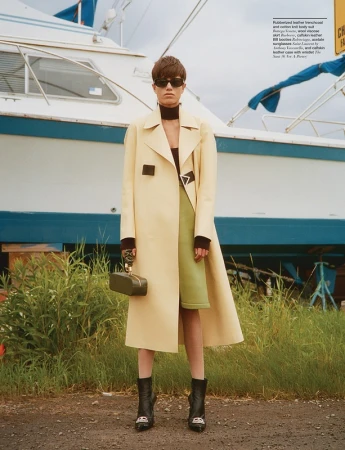 Model in cream trench coat and sunglasses holding a dark handbag in front of a white and blue boat