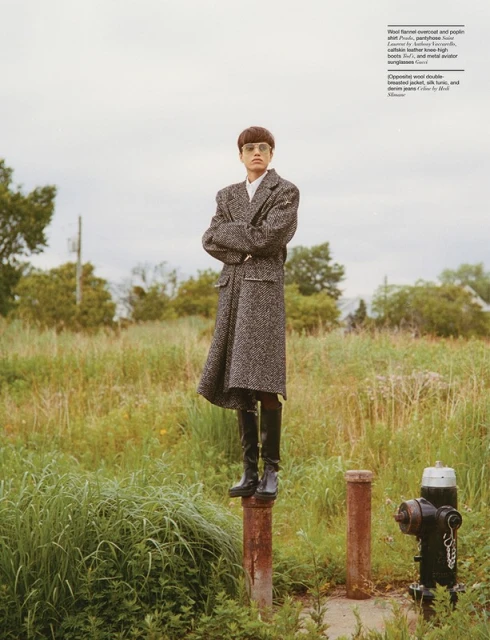 Fashion editorial shot outdoors features a model in a tweed coat and boots, standing on a metal post in a grassy field under a cloudy sky.