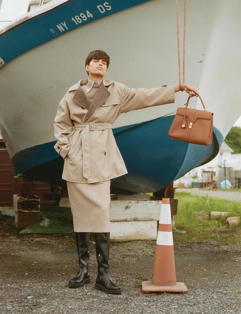 Stylish woman poses with a handbag in front of a boat, wearing a trench coat, skirt, and boots, in an outdoor setting.