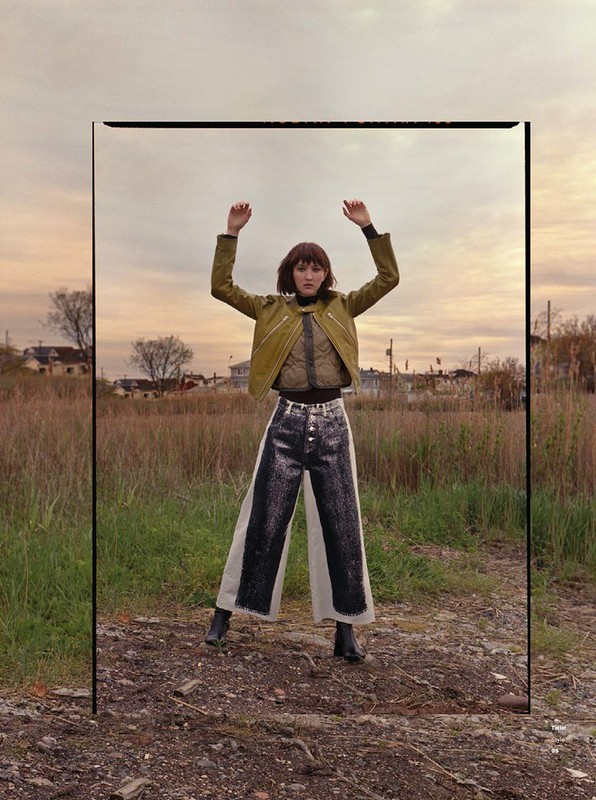 A woman stands in an open field wearing a green jacket and two-tone pants, shot through a framed perspective with a cloudy sky.