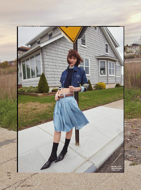 A fashion shot of a young adult female posing on a sidewalk in a denim jacket and light blue skirt in front of a house.