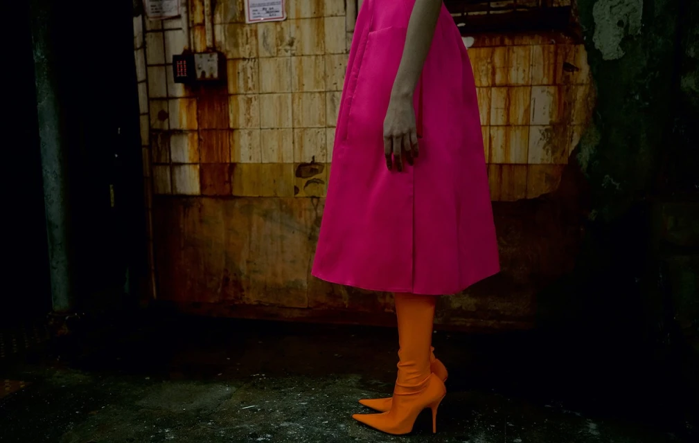 Woman's fashion shot featuring a vibrant pink dress and orange high-heeled boots against an industrial, grungy wall. 