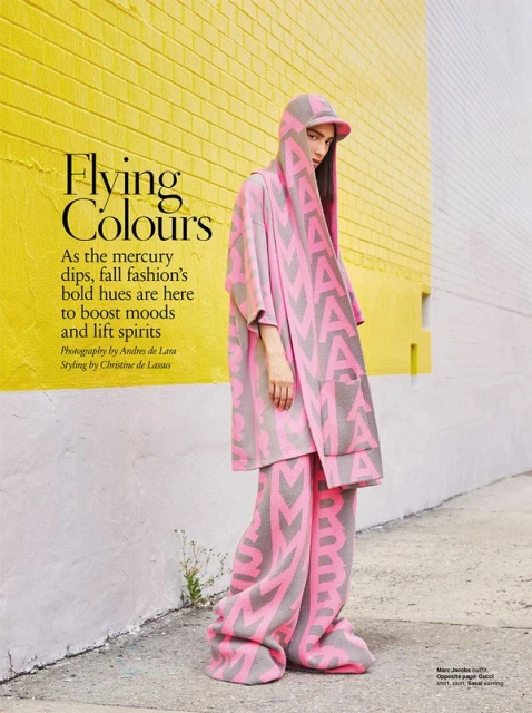 Fashion shot of a woman in a pink and grey branded hooded poncho and wide-leg pants, posing against a bright yellow and grey brick wall.