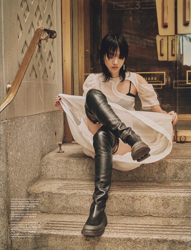 A stylish woman with blunt bangs sits on concrete steps outside, wearing a white dress, black boots, and a black bra top.