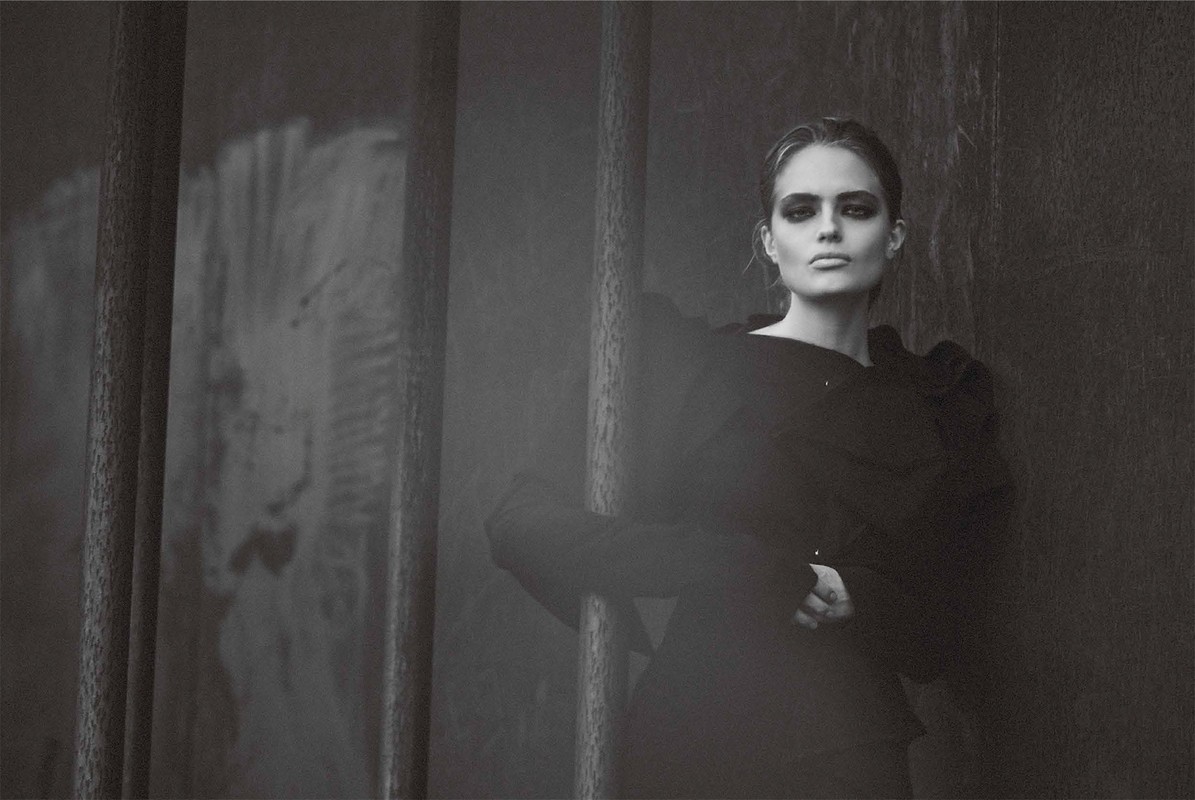 Monochrome portrait of a woman with dark eye makeup wearing a black top, posing in an indoor setting with steel bars.