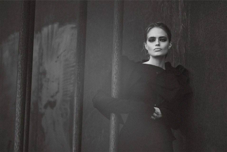 Monochrome portrait of a woman with dark eye makeup wearing a black top, posing in an indoor setting with steel bars.