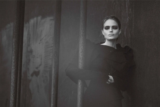 Monochrome portrait of a woman with dark eye makeup wearing a black top, posing in an indoor setting with steel bars.