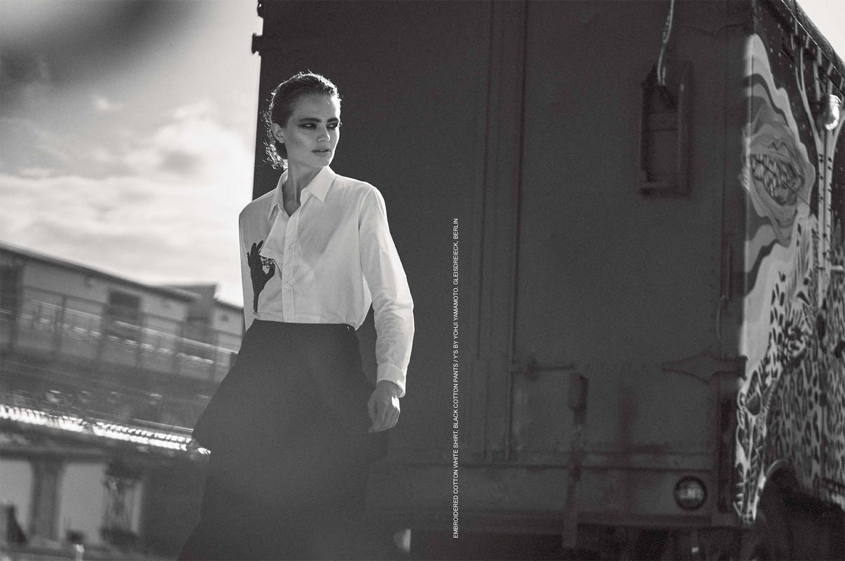 Monochrome portrait of a woman in a white shirt and black skirt outdoors with a blurred cityscape and a train carriage behind her.