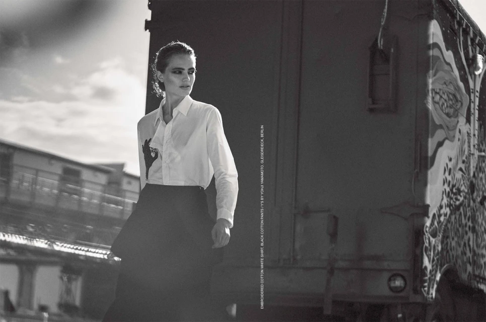 Monochrome portrait of a woman in a white shirt and black skirt outdoors with a blurred cityscape and a train carriage behind her.