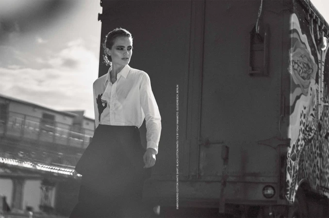Monochrome portrait of a woman in a white shirt and black skirt outdoors with a blurred cityscape and a train carriage behind her.