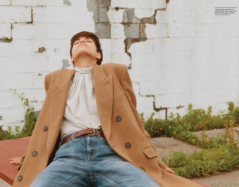 Woman in camel coat, jeans, and striped shirt sitting outside a white brick wall with green plants, looking up in sunlight.