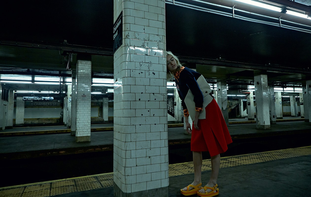 A young woman in a color-blocked outfit stands near a column in a dimly lit subway station, waiting for a train on the platform.