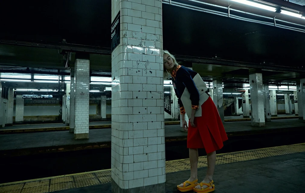 A young woman in a color-blocked outfit stands near a column in a dimly lit subway station, waiting for a train on the platform.