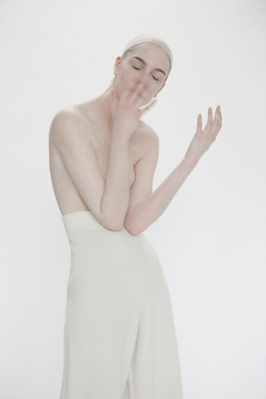 Monochromatic portrait of a person with closed eyes, pale skin, a white head covering, and cream-colored high-waisted pants against a white backdrop.