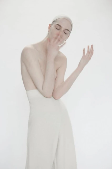 Monochromatic portrait of a person with closed eyes, pale skin, a white head covering, and cream-colored high-waisted pants against a white backdrop.
